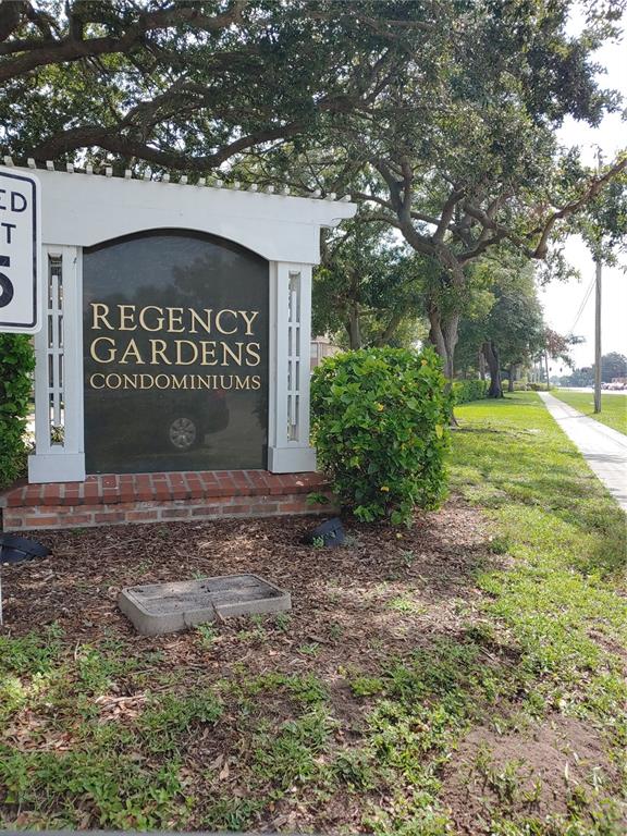 A white monument sign for Regency Gardens Condominiums stands in a landscaped area with trees — 5635 DEVONBRIAR WAY UNIT K204, ORLANDO, FL