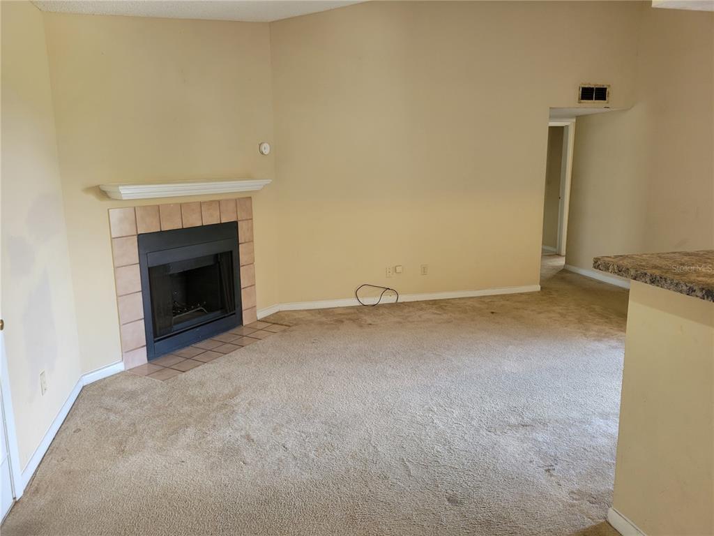 A living room features beige carpet, tan walls, and a corner fireplace with tiled surround — 5635 DEVONBRIAR WAY UNIT K204, ORLANDO, FL