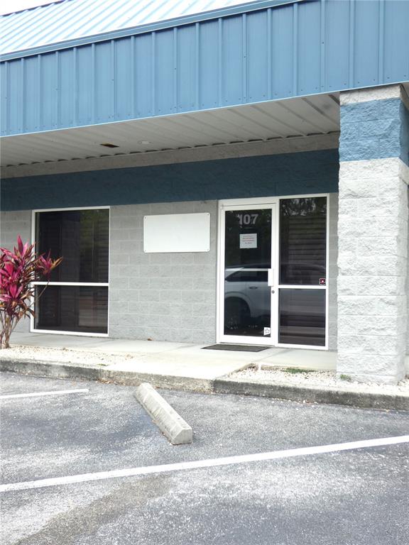 The exterior of a commercial building features a blue metal roof, gray block walls, and a glass entrance door labeled 107 — 1882 PORTER LAKE DRIVE UNIT 107/108, SARASOTA, FL