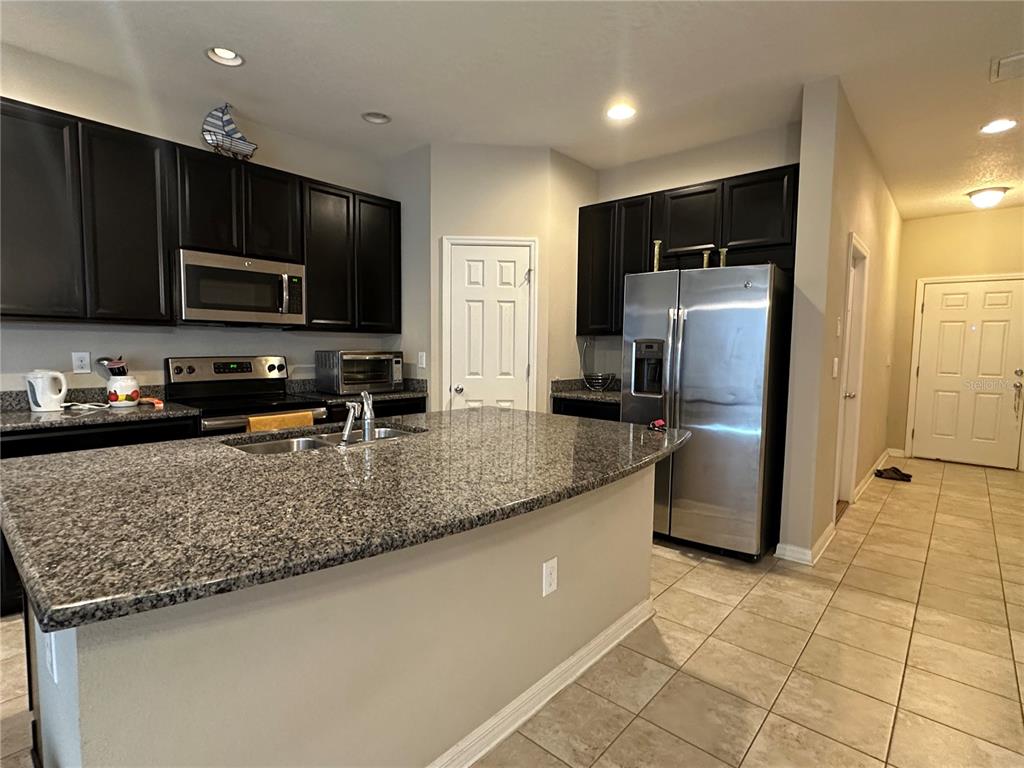 A kitchen features dark cabinets, granite countertops, a stainless steel refrigerator, and tile flooring — 7089 WOODCHASE GLEN DRIVE, RIVERVIEW, FL