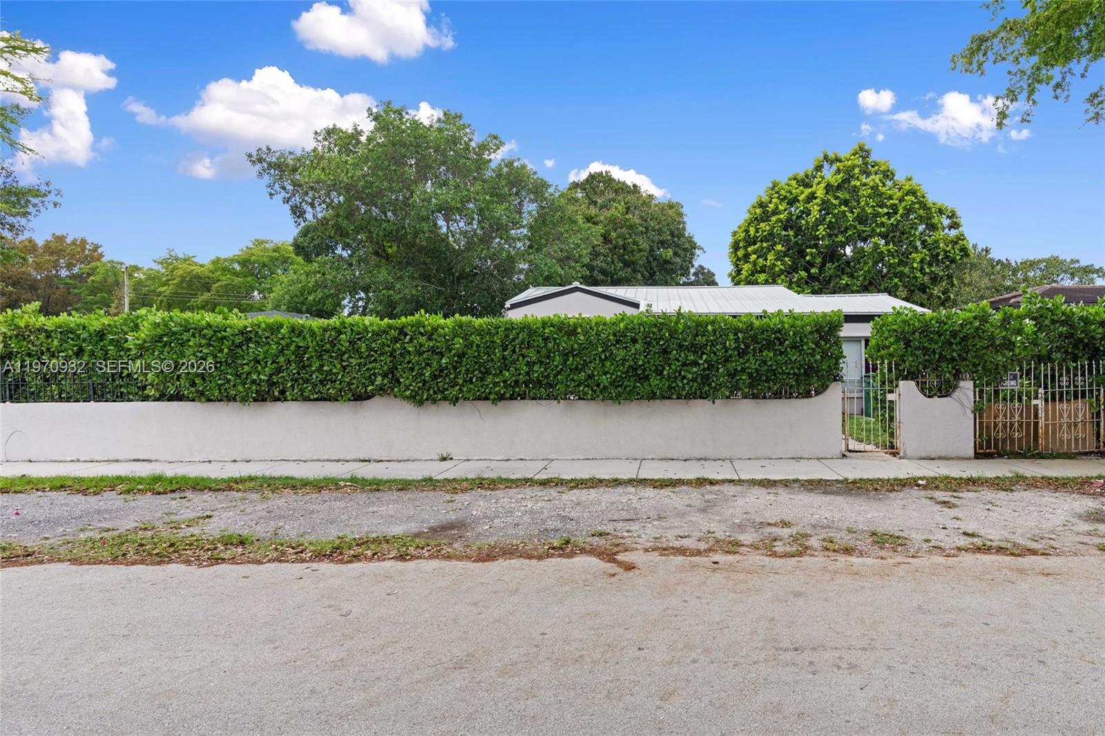 A white stucco wall and tall green hedge front a house with a metal roof — 700 SW 44th Pl, Miami FL 33134, MIAMI, FL
