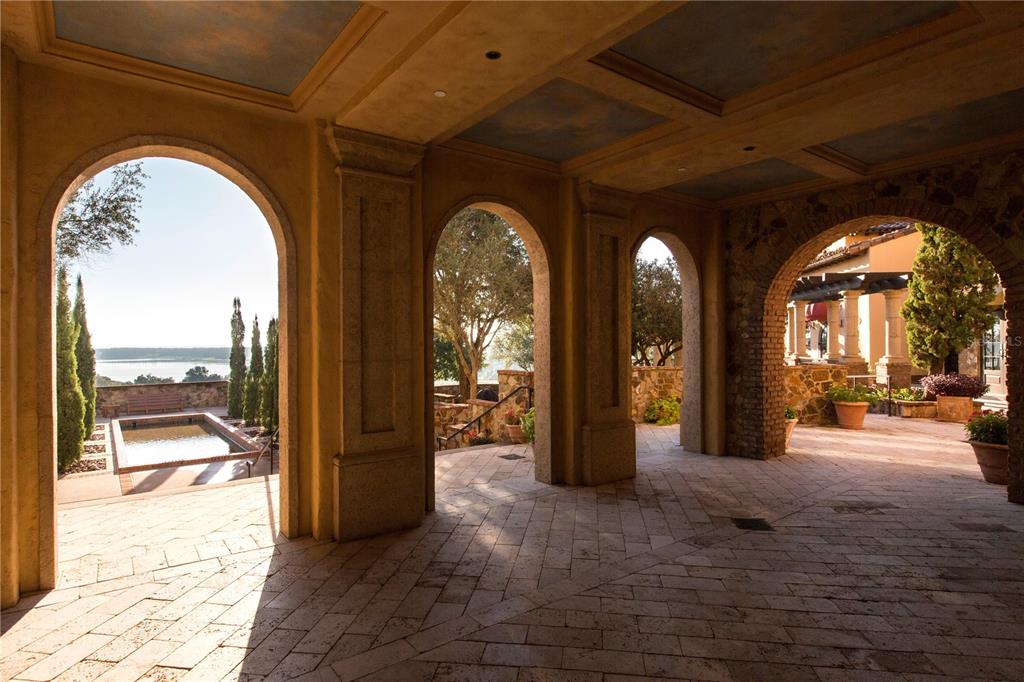 A covered stone patio features multiple arched openings overlooking a pool and a distant lake — 17114 MEDICI WAY, BELLA COLLINA, FL