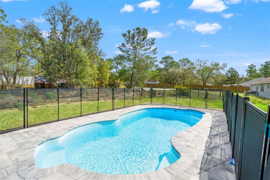 A backyard swimming pool with a safety fence surrounded by a paved deck and trees — 12015 PHOENIX AVENUE, WEEKI WACHEE, FL