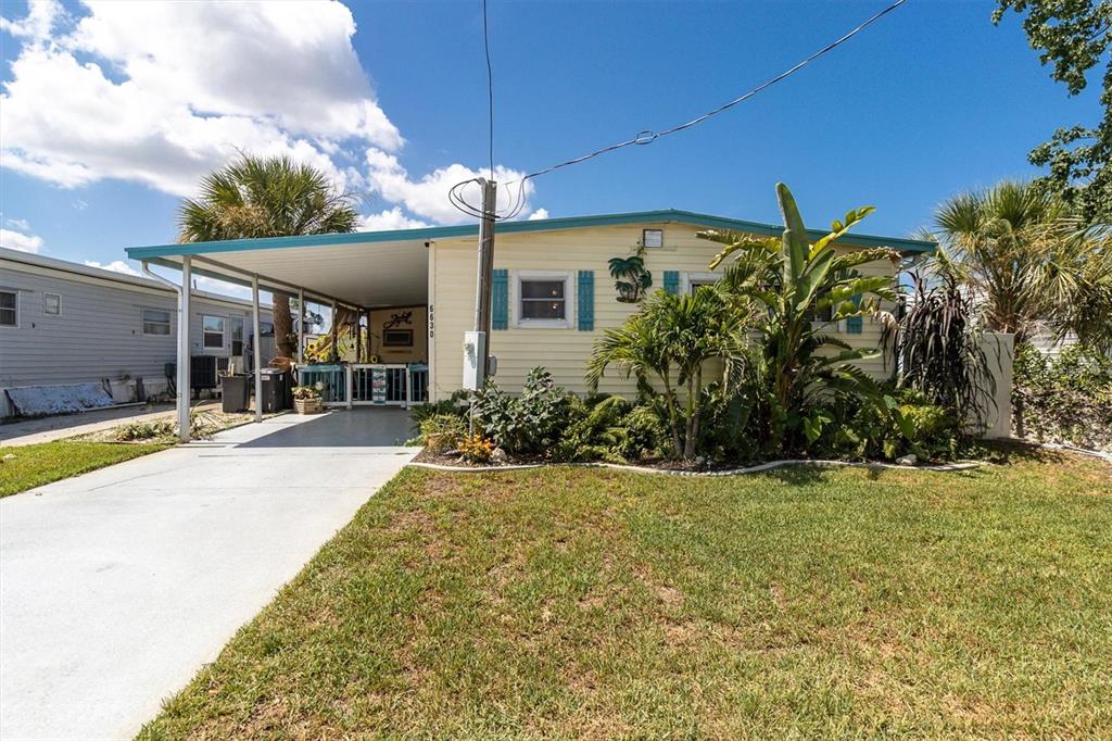 A light yellow manufactured home features a carport, front lawn, and tropical landscaping — 6630 TOWER DRIVE, HUDSON, FL