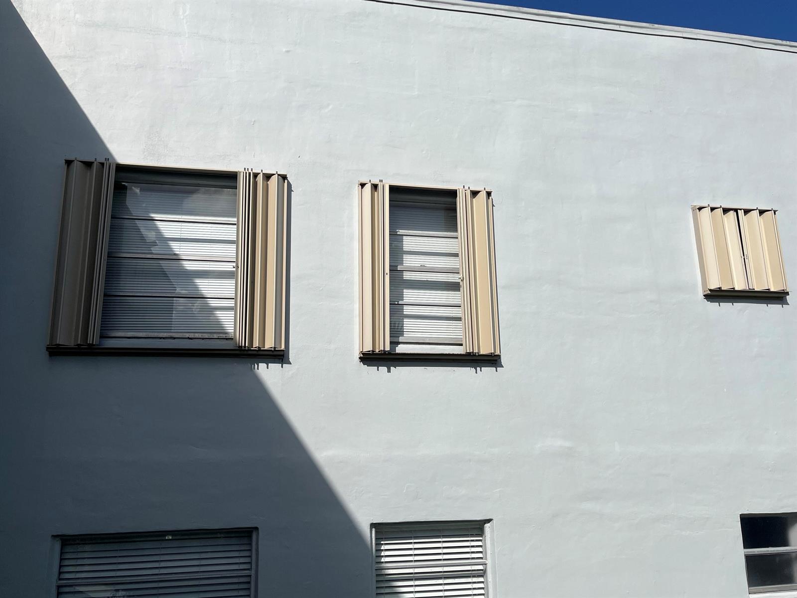 A light-colored exterior wall features several windows with beige accordion-style hurricane shutters — 142 Normandy C, Delray Beach FL 33484, DELRAY BEACH, FL