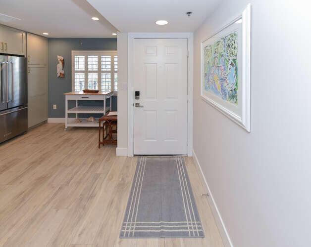 A hallway with light wood flooring leads to a white front door and kitchen area — 275 Palm Avenue # C406, Jupiter FL 33477, JUPITER, FL