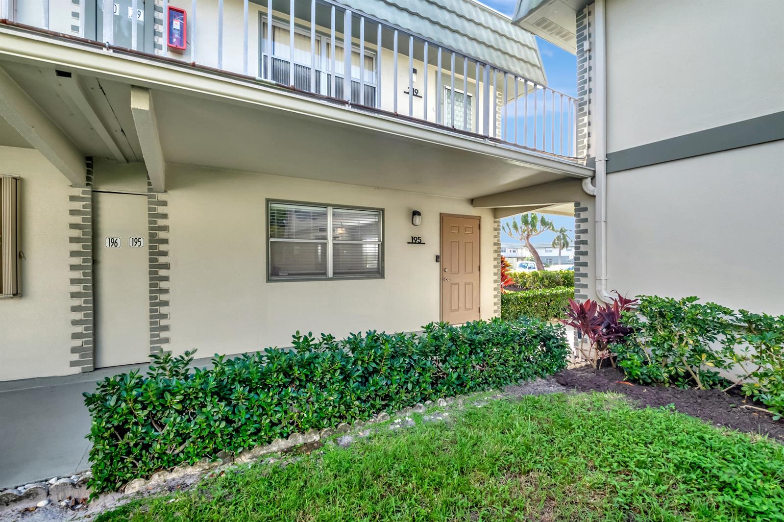 The exterior of a two-story apartment building features a tan door, window, and green landscaping — 195 Monaco E # 195, Delray Beach FL 33446, DELRAY BEACH, FL