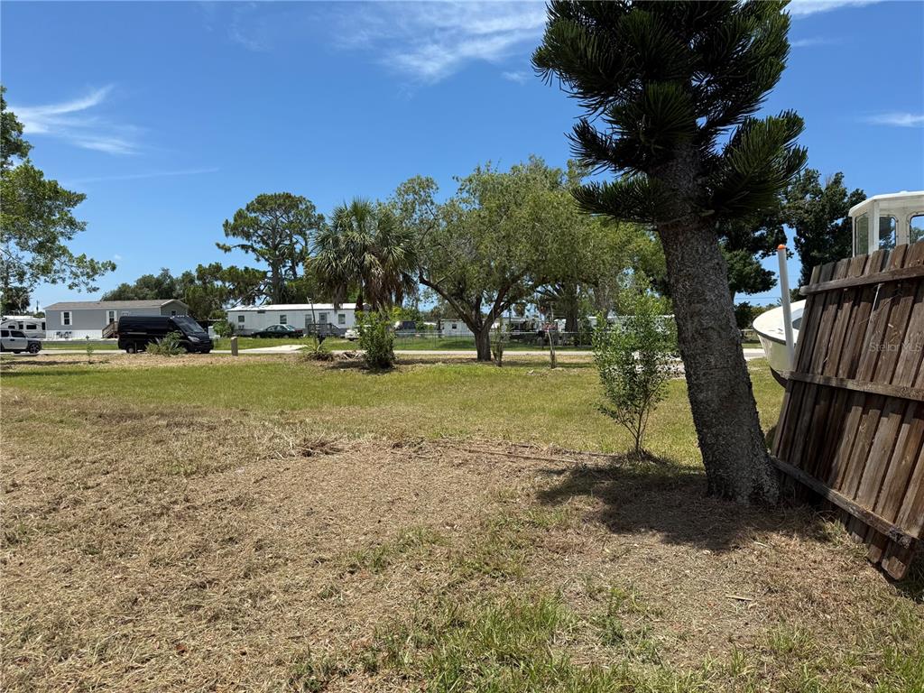 A grassy lot features a large tree, a wooden fence, and a boat in the background — 3825 BRAVO ROAD, PORT CHARLOTTE, FL