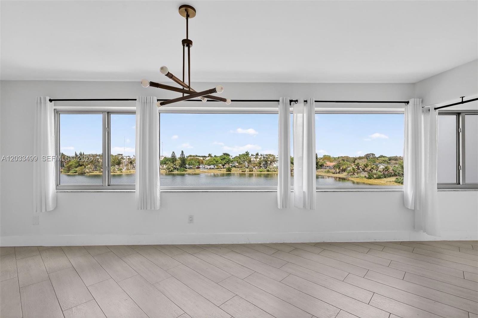 A bright room features light wood-look tile floors, a modern chandelier, and large lake-view windows — 877 NE 195th St # 422, Miami FL 33179, MIAMI, FL
