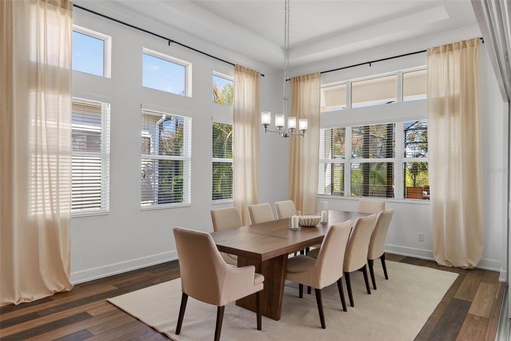 A dining room features a wooden table, upholstered chairs, tall windows, and light-colored curtains — 8276 REDONDA LOOP, LAKEWOOD RANCH, FL