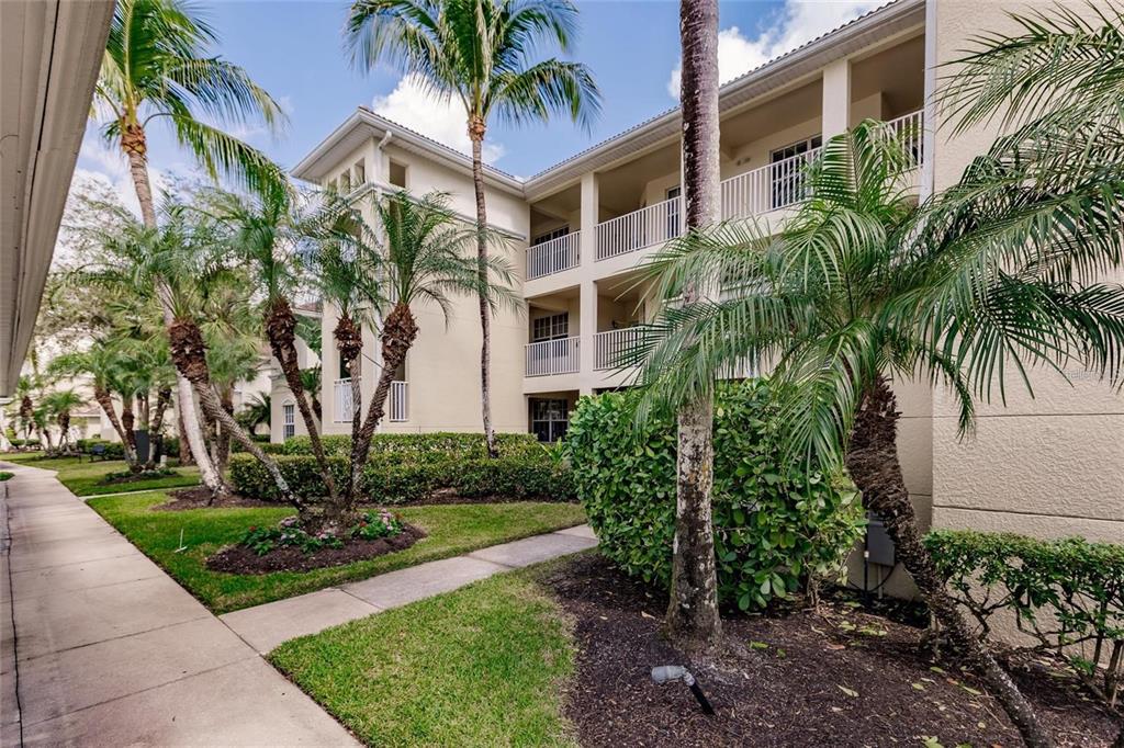 A sidewalk leads past palm trees and landscaping toward a multi-story tan apartment building — 1878 TARPON BAY DRIVE S UNIT 102, NAPLES, FL