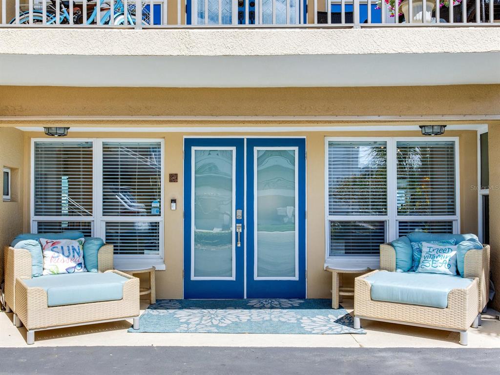A covered patio features two wicker chairs flanking open French doors leading into a living room — 210 DOLPHIN POINT UNIT B, CLEARWATER BEACH, FL