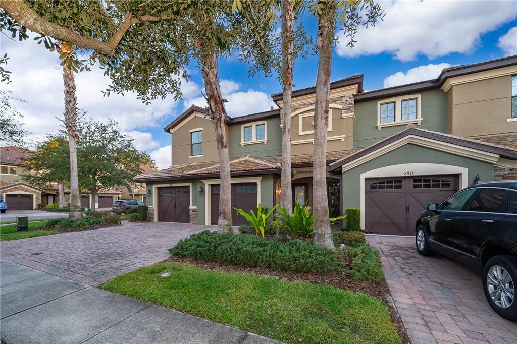 A multi-story townhouse exterior features a brick driveway, three garage doors, and palm trees — 8945 AZALEA SANDS LANE UNIT 8945, CHAMPIONS GATE, FL