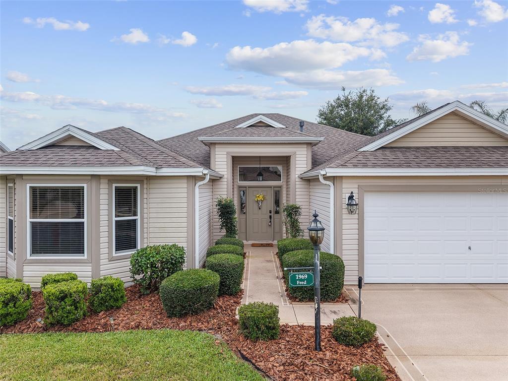 A single-story house with tan siding, a front-facing garage, and a landscaped walkway — 1969 HAGOOD LOOP, THE VILLAGES, FL