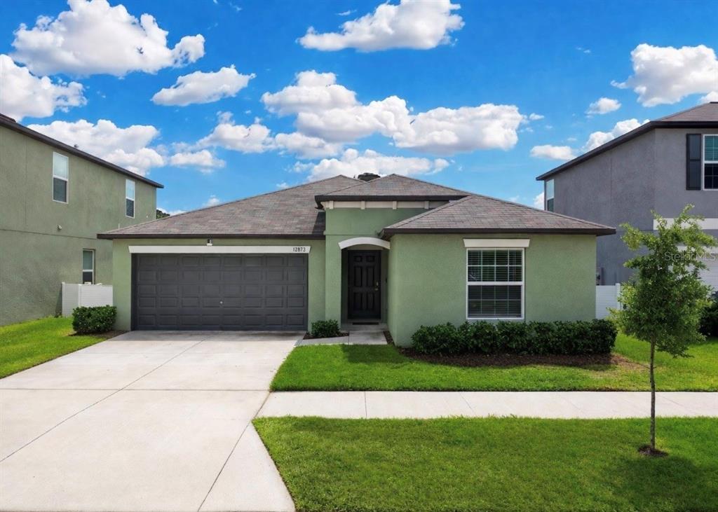 A single-story green stucco house with a two-car garage and a front lawn — 12873 TRIPOLI AVENUE, RIVERVIEW, FL