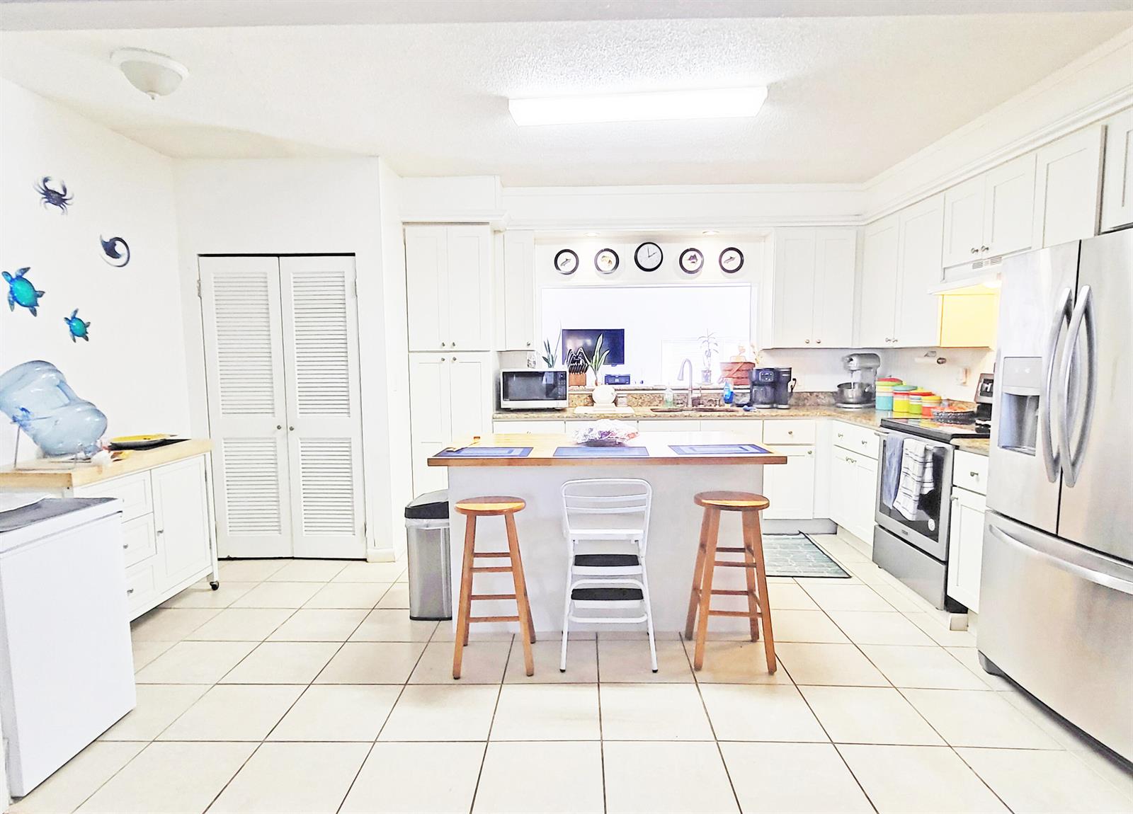 A bright kitchen features white cabinetry, a central island with three stools, and tile flooring — 203 Melton Drive, Fort Pierce FL 34982, FORT PIERCE, FL