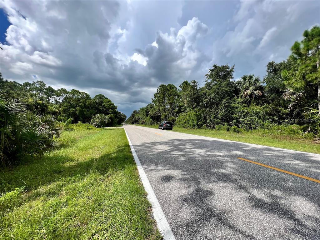 A paved road stretches into the distance, flanked by dense green trees and grassy shoulders — 168 PRINEVILLE STREET, PORT CHARLOTTE, FL