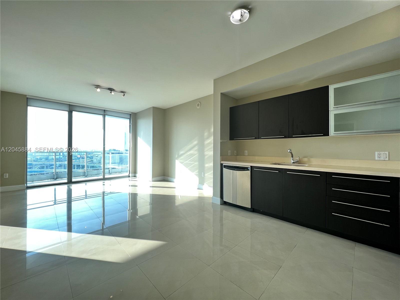 A modern kitchen with dark cabinetry and stainless steel appliances opens into a bright living area — 90 SW 3rd St # 1710, Miami FL 33130, MIAMI, FL