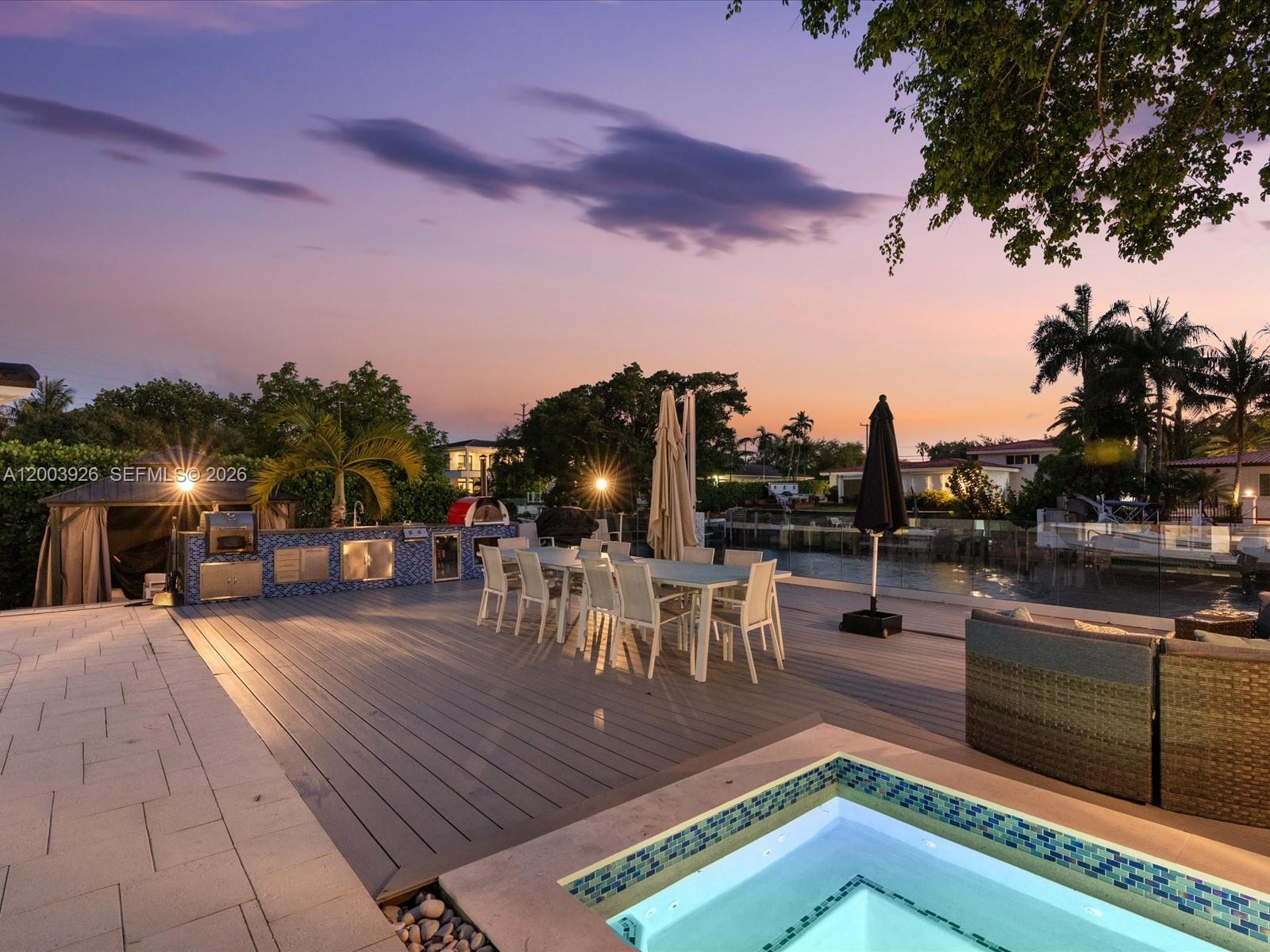 An outdoor deck features a dining set, a built-in kitchen, and a pool at sunset — 5000 Riviera Dr, Coral Gables FL 33146, CORAL GABLES, FL