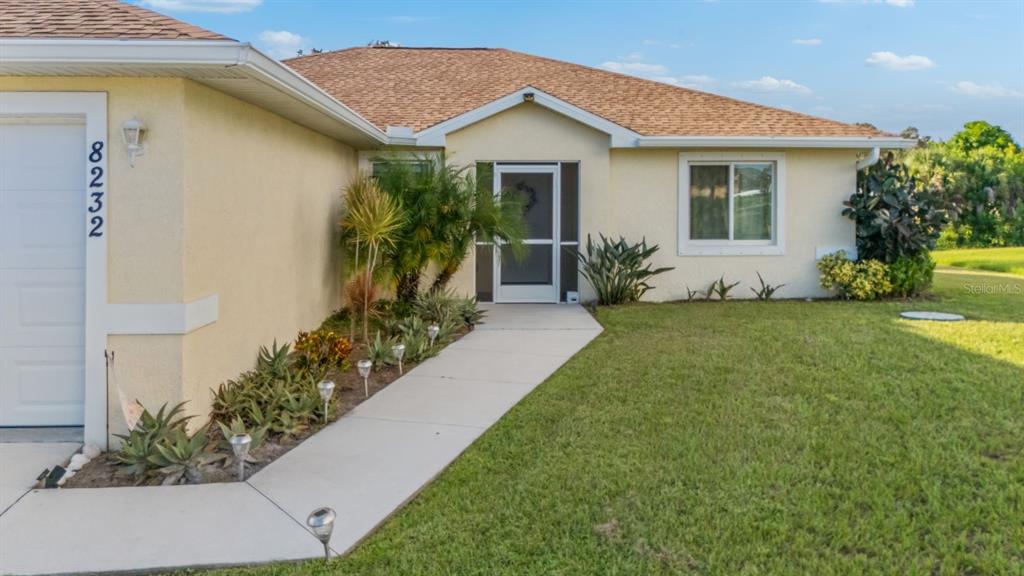 The exterior of a single-story house features a tan stucco facade, a walkway, and landscaping — 8232 WAWANA ROAD, NORTH PORT, FL