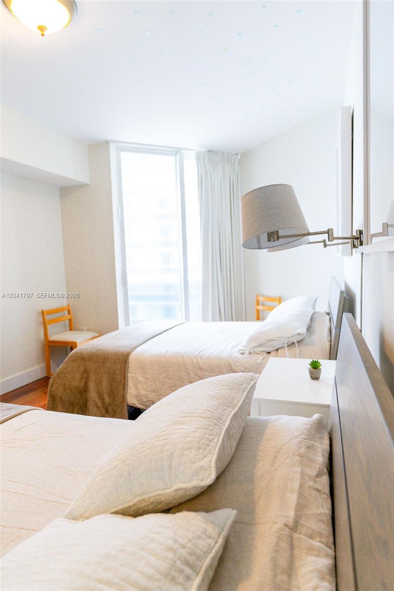 A bedroom features two beds, a wall-mounted lamp, and a window with white curtains — 9595 Collins Ave # C, Surfside FL 33154, SURFSIDE, FL