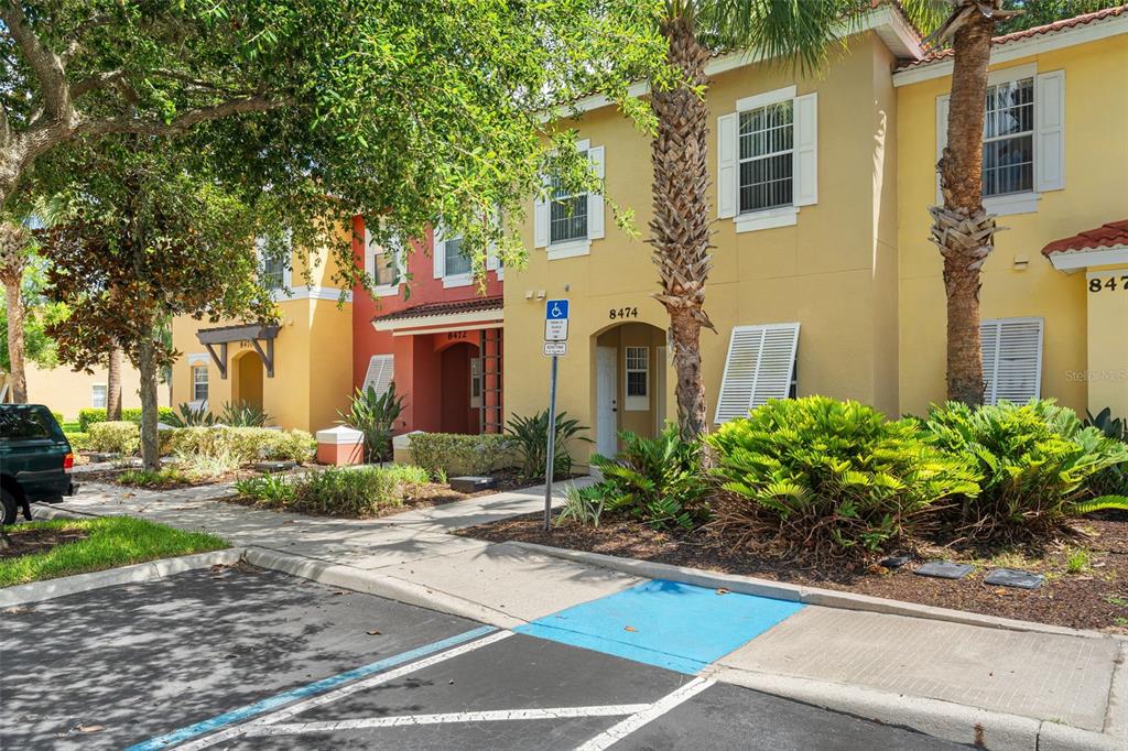 A multi-story townhouse complex features yellow and red stucco facades with an accessible parking space — 8474 CRYSTAL COVE LOOP, KISSIMMEE, FL