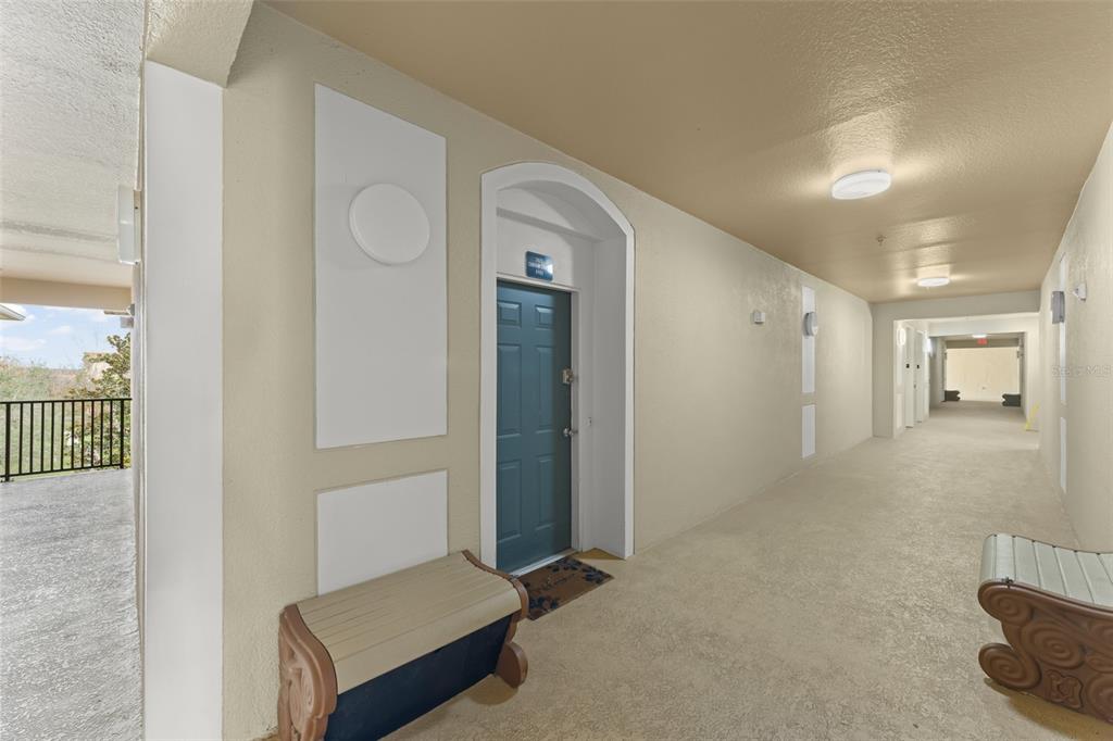 An outdoor apartment hallway features a blue door, a bench, and beige walls — 7675 COMROW STREET UNIT 405, KISSIMMEE, FL