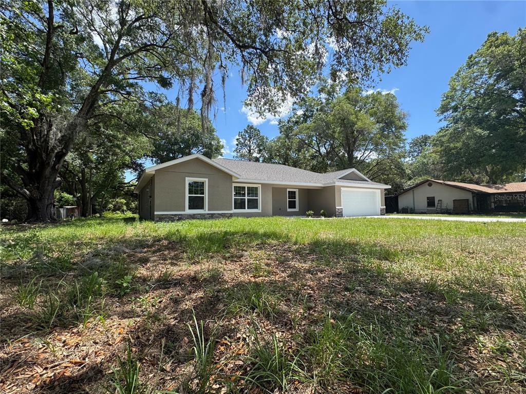 A tan, single-story house with a two-car garage sits on a grassy, tree-filled lot — 8121 SW 202ND TERRACE, DUNNELLON, FL