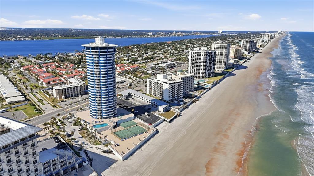 An aerial view shows a tall, cylindrical blue-and-white high-rise building situated on a sandy beach — 2625 S ATLANTIC AVENUE UNIT 18NW, DAYTONA BEACH, FL