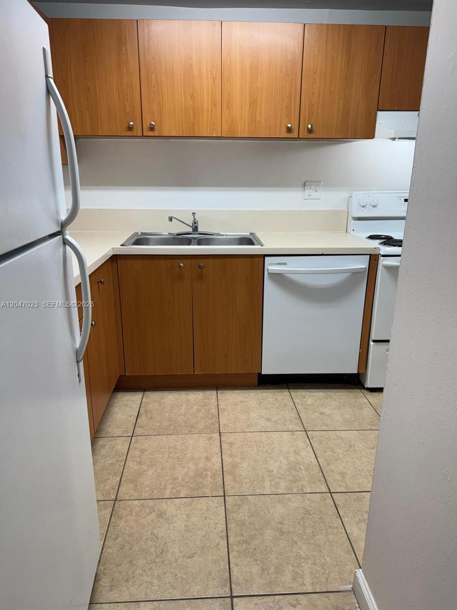 A kitchen features wood-tone cabinets, white appliances, a double sink, and beige tile flooring — 411 Executive Center Dr # 203, West Palm Beach FL 33401, WEST PALM BEACH, FL