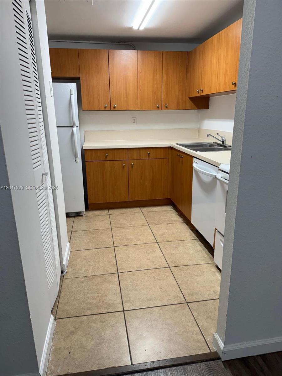 A galley kitchen features wood cabinets, white appliances, and beige tile flooring — 411 Executive Center Dr # 203, West Palm Beach FL 33401, WEST PALM BEACH, FL