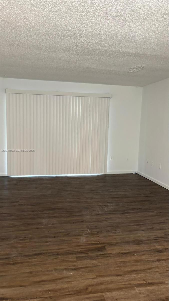 An empty room features dark wood-look flooring, white walls, and vertical blinds covering a sliding door — 411 Executive Center Dr # 203, West Palm Beach FL 33401, WEST PALM BEACH, FL