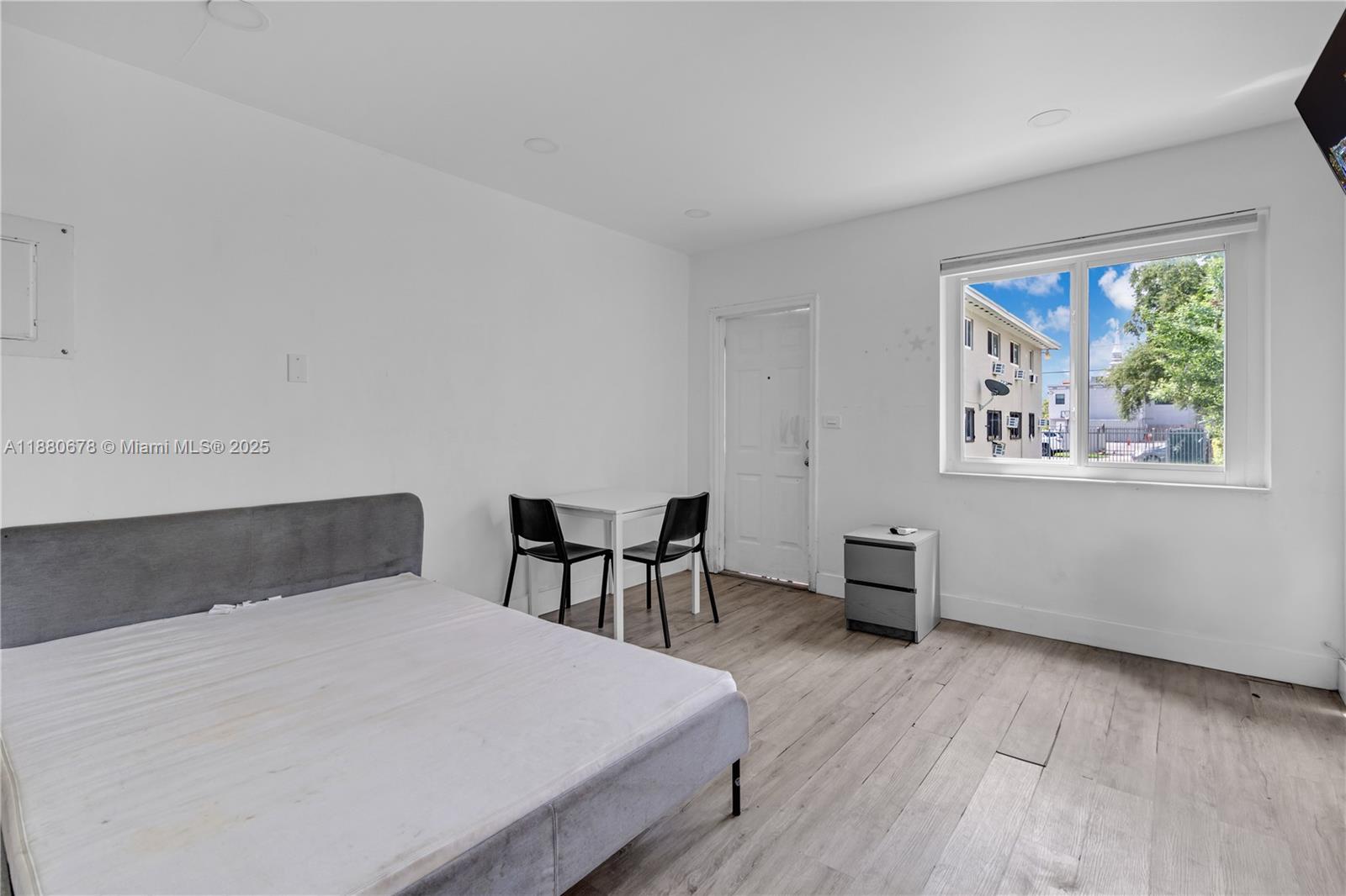 A bedroom features white walls, light wood-look flooring, a gray bed, and a small dining set — 110 NW 9th Ave # 3, Miami FL 33128, MIAMI, FL