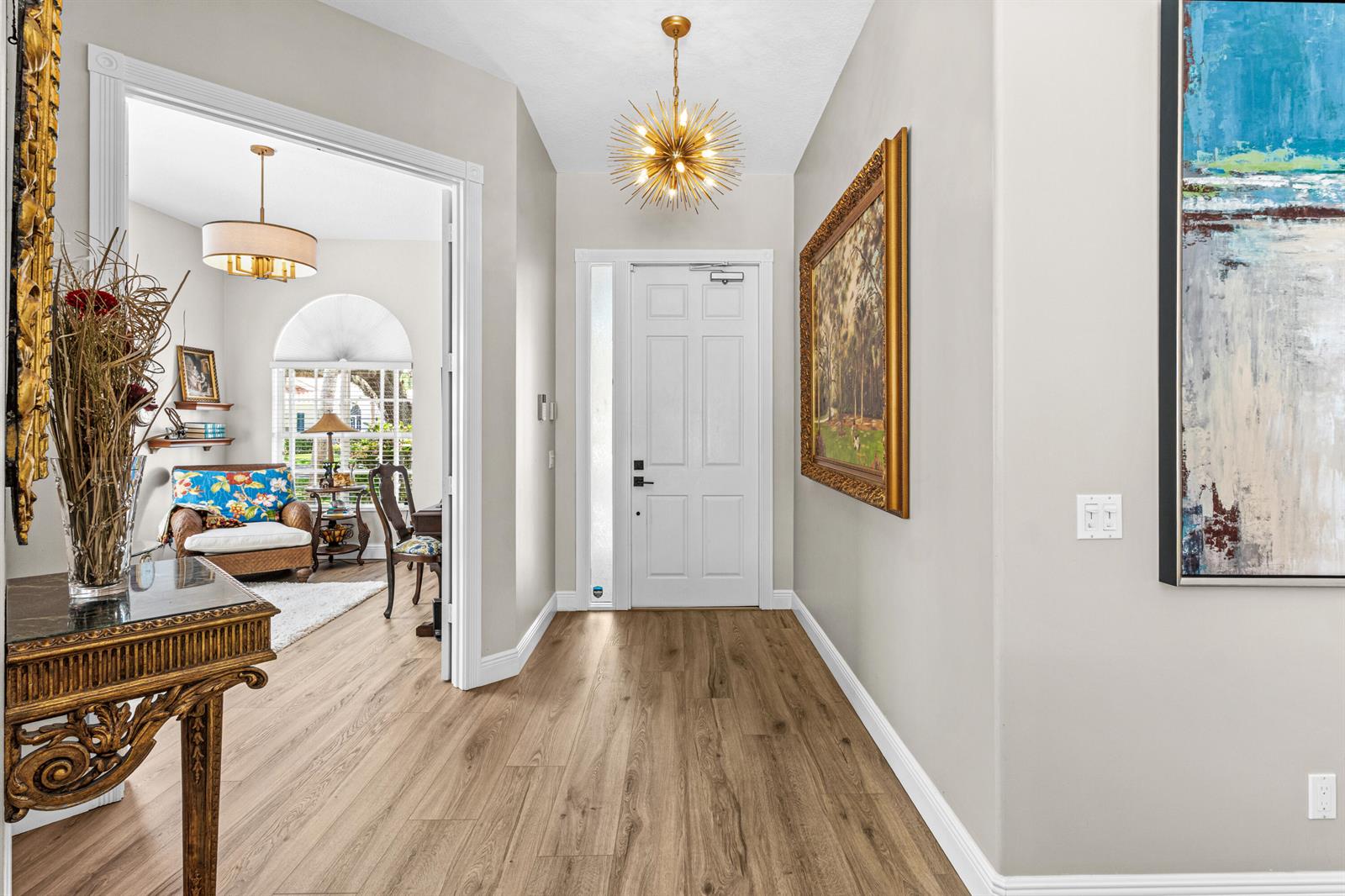A bright entryway features light wood flooring, a gold starburst chandelier, and framed wall art — 4983 Garden Drive, Delray Beach FL 33445, DELRAY BEACH, FL