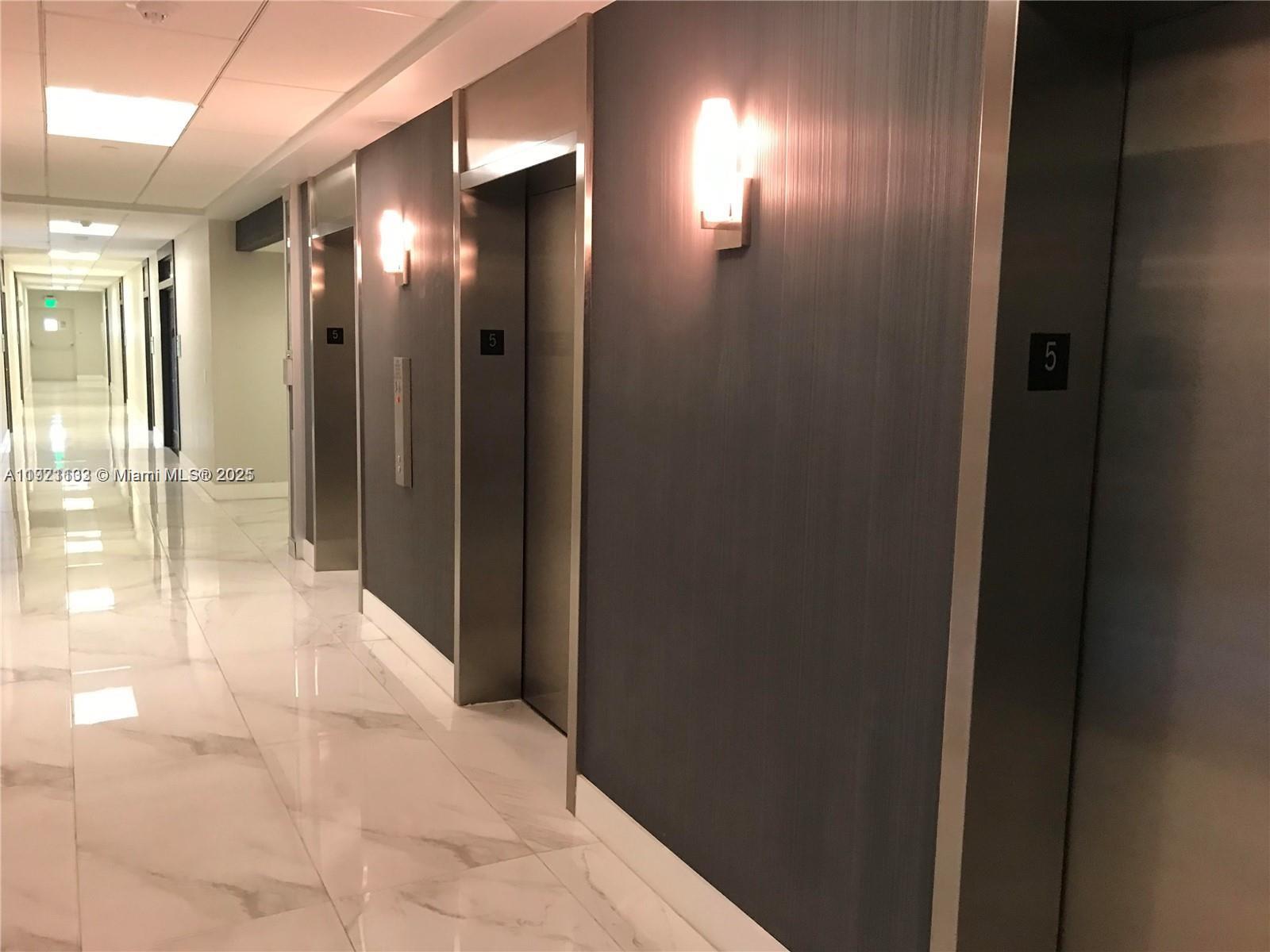 A hallway features marble floors and three stainless steel elevator doors with dark wall paneling — 150 SE 2nd Ave, Miami FL 33131, MIAMI, FL
