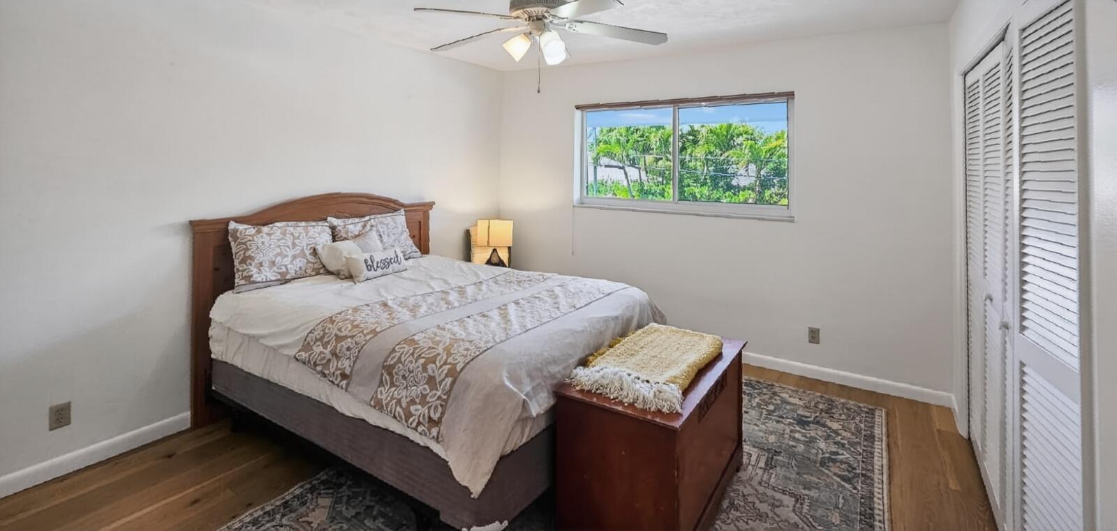 A bedroom features a wooden bed frame, a storage chest, and a window with palm trees — 8 Garden Street # 206r, Tequesta FL 33469, TEQUESTA, FL