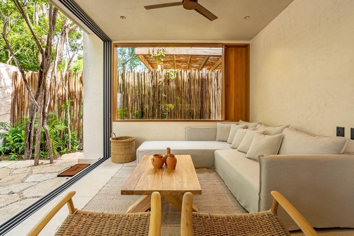 A living room with a sectional sofa, wooden coffee table, and large window overlooking bamboo fencing — Amari Tulum 10 Avenida Norte & Calle Palma, FL