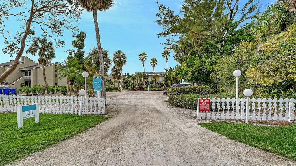 A dirt driveway leads toward apartment buildings framed by palm trees and white picket fences — 5311 GULF OF MEXICO DRIVE UNIT 5, LONGBOAT KEY, FL