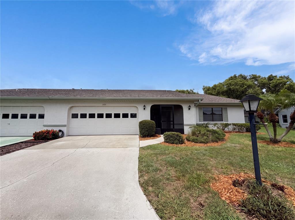 A single-story house exterior features a white stucco facade, a two-car garage, and a lawn — 1404 LELAND DRIVE UNIT 0, SUN CITY CENTER, FL
