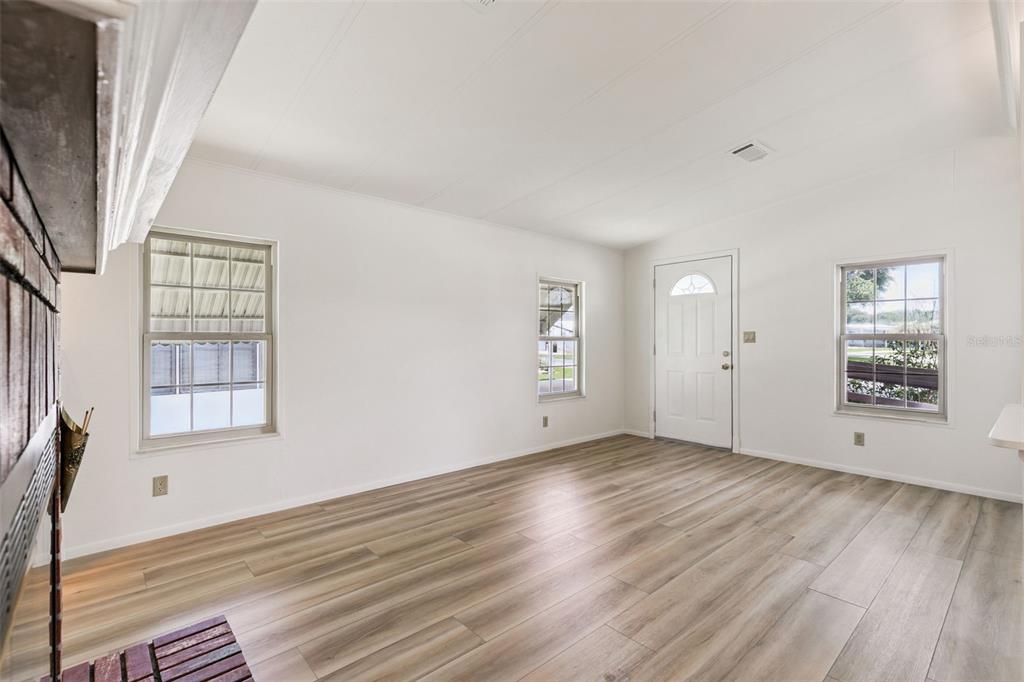 An empty living room with white walls, wood-look flooring, a brick fireplace, and three windows — 938 BLUE HERON BOULEVARD, OSTEEN, FL