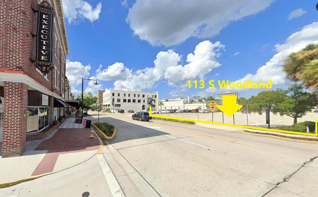 A vacant, fenced-in dirt lot sits next to a brick building on a city street — 113 S WOODLAND BOULEVARD, DELAND, FL