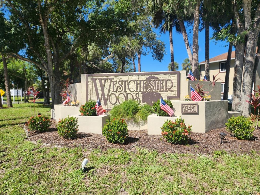A beige community entrance sign for Westchester Woods stands surrounded by landscaping and American flags — 22481 WESTCHESTER BOULEVARD UNIT A19, PUNTA GORDA, FL
