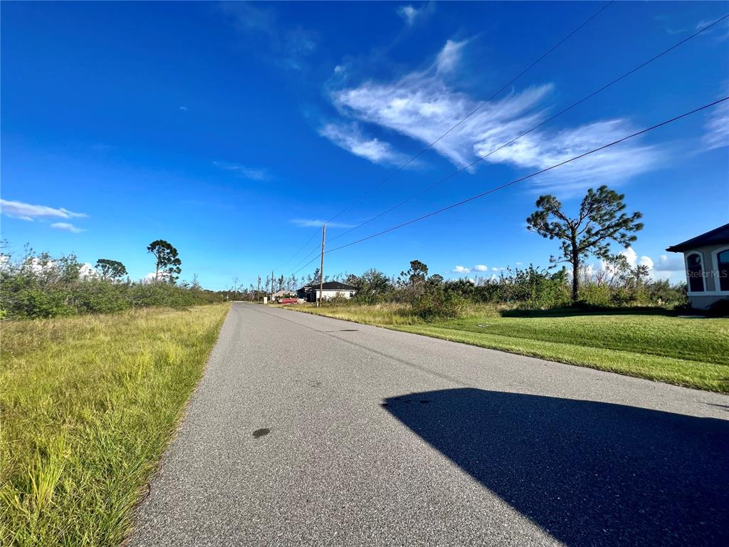 A paved road stretches into the distance past grassy lots and scattered trees under a sky — 15755 STAUNTON CIRCLE, PORT CHARLOTTE, FL