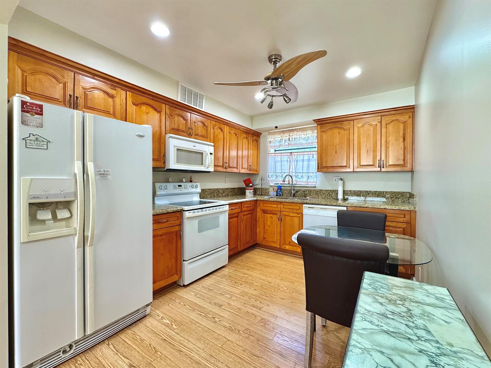 A kitchen features wood cabinets, granite countertops, white appliances, and a small glass dining table — 565 Piedmont L, Delray Beach FL 33484, DELRAY BEACH, FL