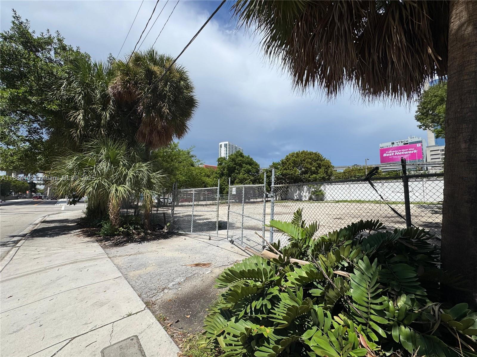 A fenced, vacant dirt lot is bordered by a sidewalk, palm trees, and lush greenery — 201 SE 2nd Ave, Miami FL 33131, MIAMI, FL