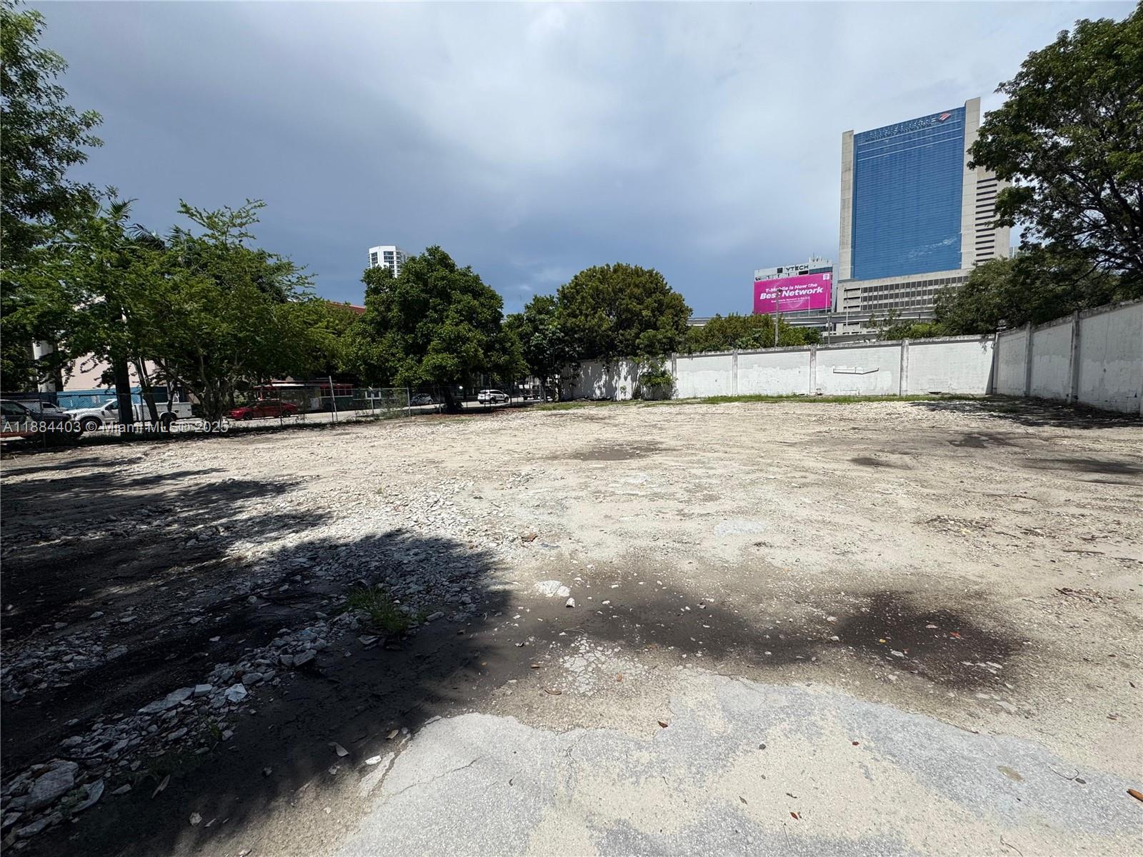 A large, vacant, unpaved urban lot is enclosed by a white wall and surrounding trees — 201 SE 2nd Ave, Miami FL 33131, MIAMI, FL