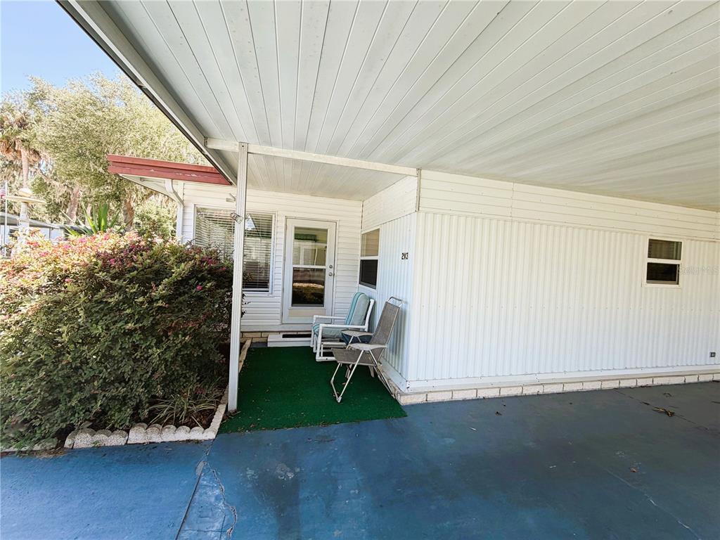 A mobile home exterior features a covered carport, a small green patio, and two chairs — 203 ASPEN CIRCLE, LEESBURG, FL
