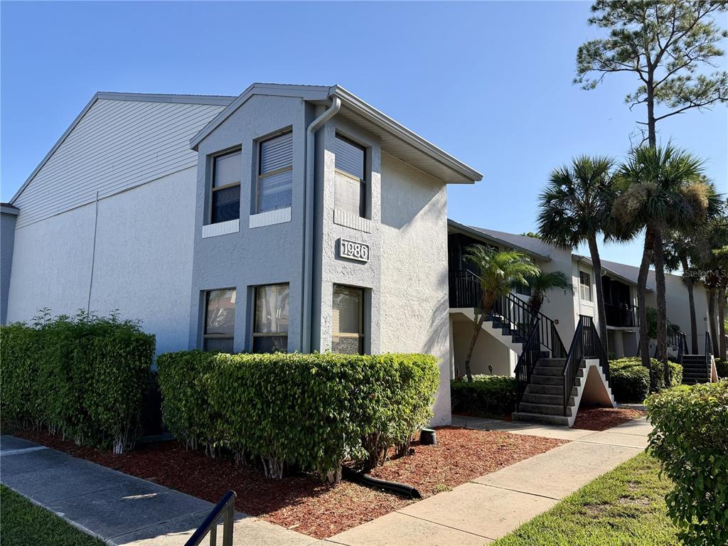 A two-story apartment building exterior features light-colored stucco walls, dark-framed windows, and an outdoor staircase — 1986 PELICAN LANDING BOULEVARD UNIT 1511, CLEARWATER, FL