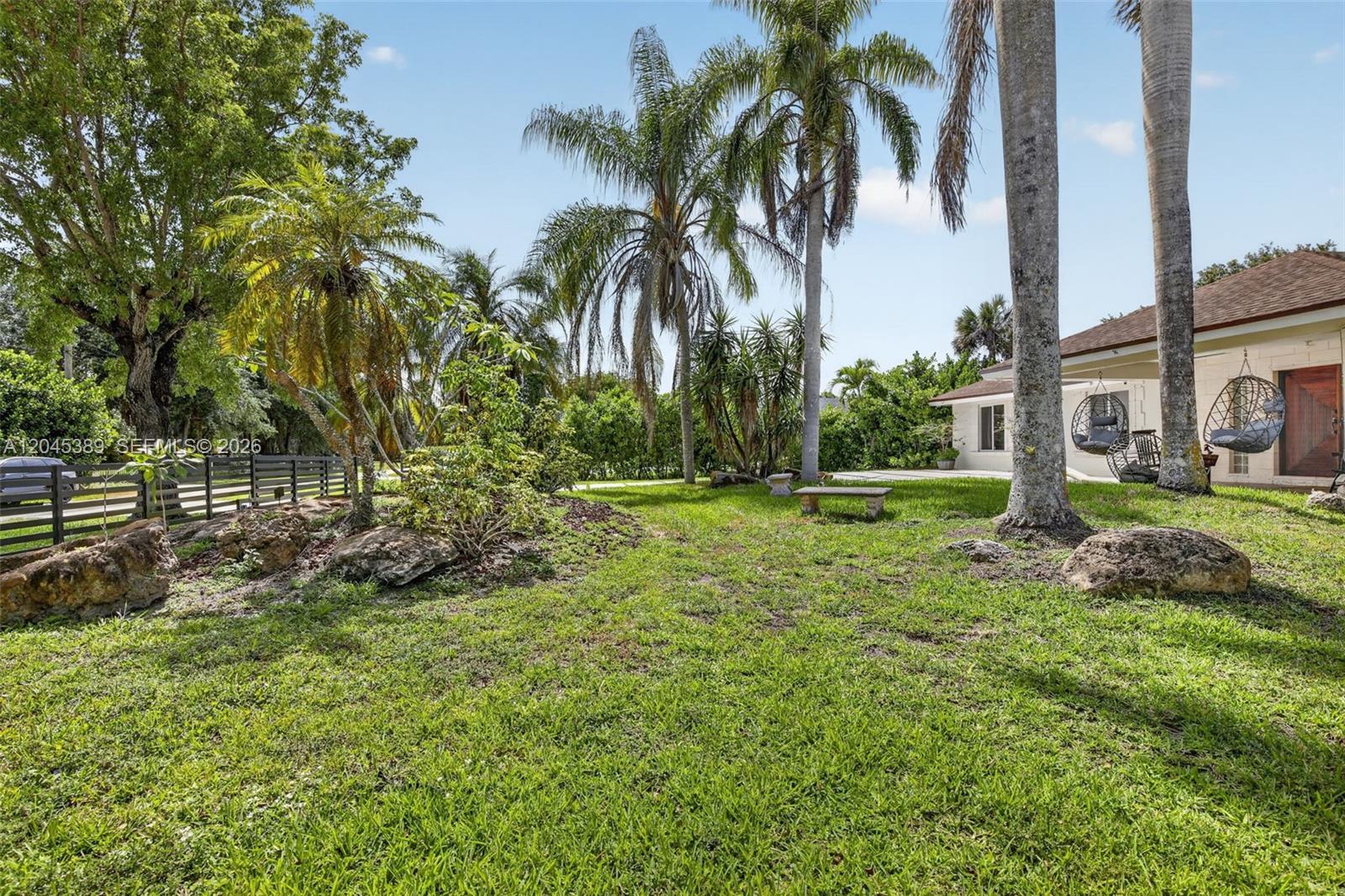 A grassy backyard features palm trees, large decorative rocks, and hanging chairs near a house — 14401 SW 23rd St, Davie FL 33325, DAVIE, FL