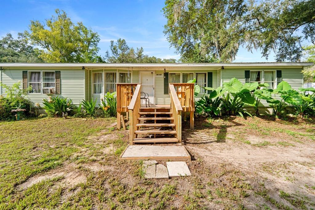 A light green single-story house features a wooden front porch with stairs and large plants — 16143 PALACKY STREET, BROOKSVILLE, FL